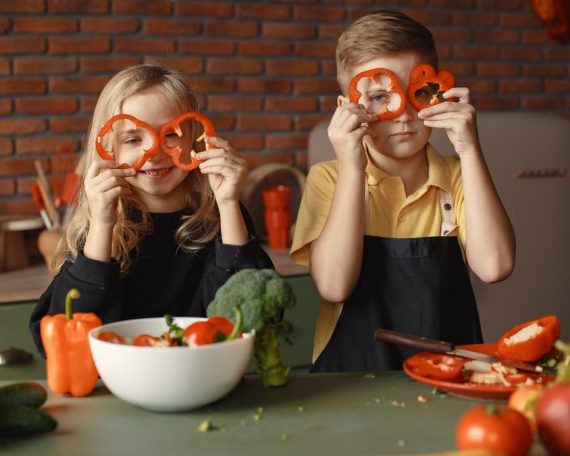 Funny boy and girl wearing apron looking through slices of pepper while preparing healthy vegetarian salad together at table with ingredients and bowls