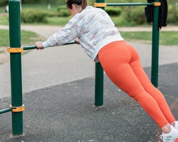 A woman in sportswear performs exercises on outdoor fitness equipment.